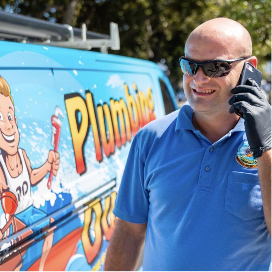 Plumbing Dudes mascot at the beach with surfboard, wrench, and toolbox